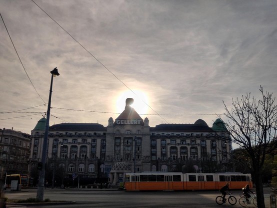 Danubio Hotel Géllert en la puesta de sol. El sol se pone detrás del torreón de la fachada y la deja en sombras. Por delante del hotel pasa un tranvía amarillo. A la derecha, dos personas pasean en bicicleta. En el cielo hay unas leves nubes y  se ven los cables del tranvía dibujados sobre ellas.