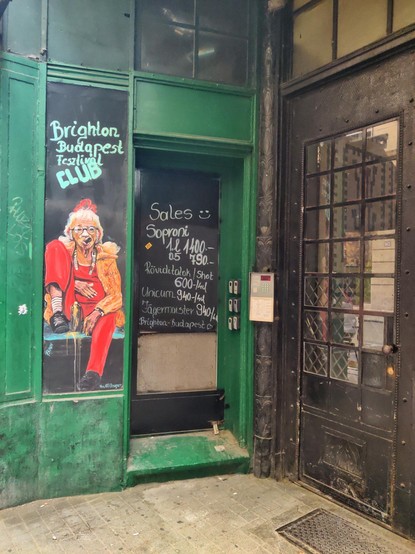 Esquina de una calle en el centro de Budapest. A la izquierda se ve la fachada de un bar pintada de verde. Hay un dibujo de una mujer de cierta edad vestida de rojo y fumando un puro. Encima hay un texto anunciando el Brighton Budapest Festival Club. A la derecha del dibujo hay un cartel indicando los precios de las bebidas. Más a la izquierda hay una puerta de hierro negra que da entrada a una vivienda