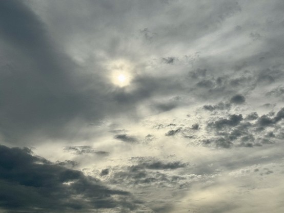 Landscape orientation, photo of the sun partially obscured by a cloud filled sky. The sun is almost mid frame and gently radiates light. Lower frame wispy gaseous, dark gray clouds drift about below a lighter gray wall cloud. The sun and gaseous dark clouds resemble a deep space nebula or whatnot.
