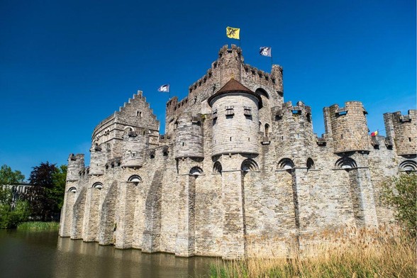 A medieval stone castle with multiple towers and turrets surrounded by a moat. The Gravensteen Castle in Ghent features light gray stonework, flying colorful flags, and is photographed on a clear day with bright blue skies. Reeds grow along the water's edge in the foreground.