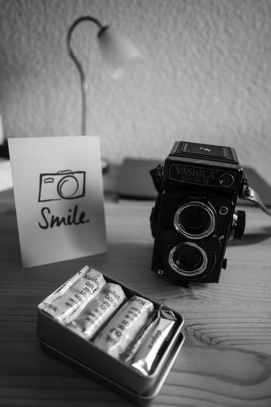 Das schwarzweiss Foto zeigt eine schwarze analoge Kamera (eine sogenannte TLR von Yashica) davor eine Box mit diversen Filmen.
Links steht eine Karte auf der "smile" steht und eine Kamera abgedruckt ist. Im Hintergrund steht eine Lampe.